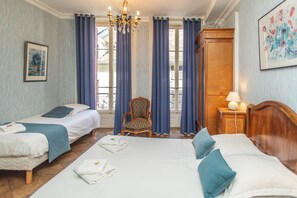 Ein Hotelzimmer mit zwei Betten, einem Sessel, einem Nachttisch, einer Chandelierleuchte und einem Fenster mit Vorhängen.