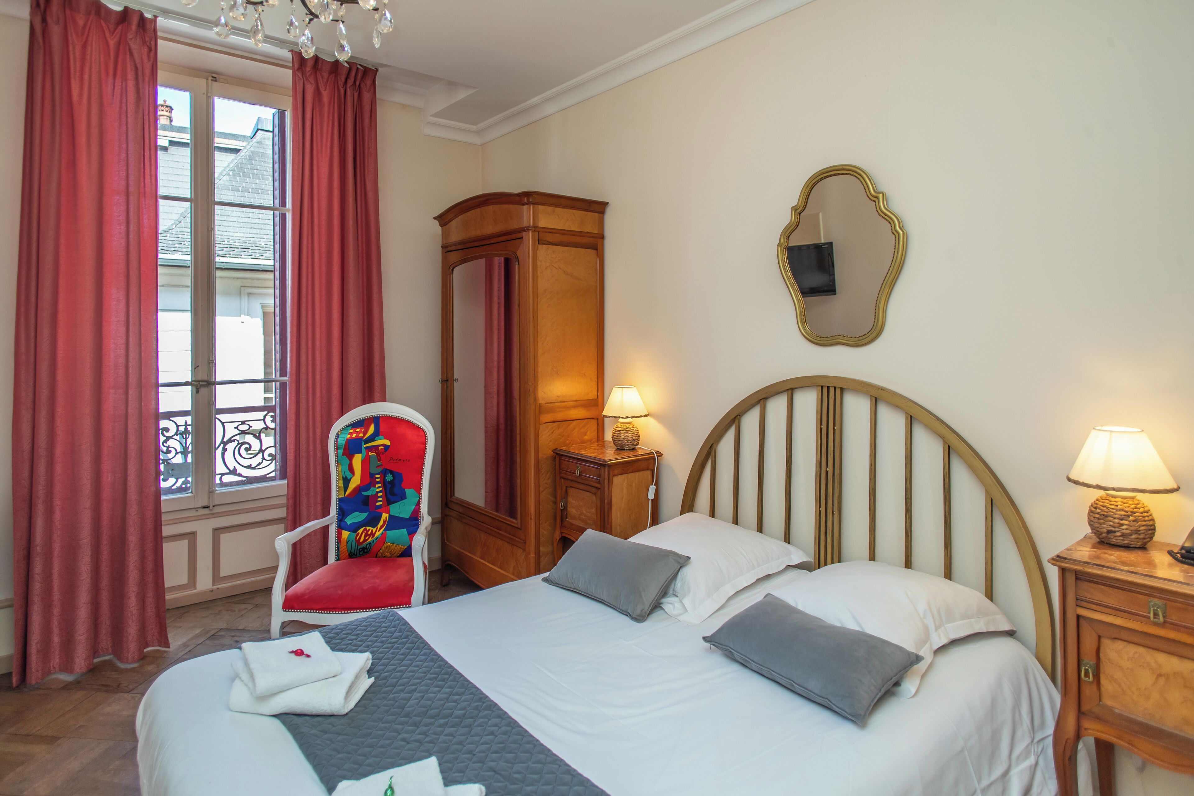 A hotel room with a bed, a chair, a wardrobe, a mirror, and a window with curtains.