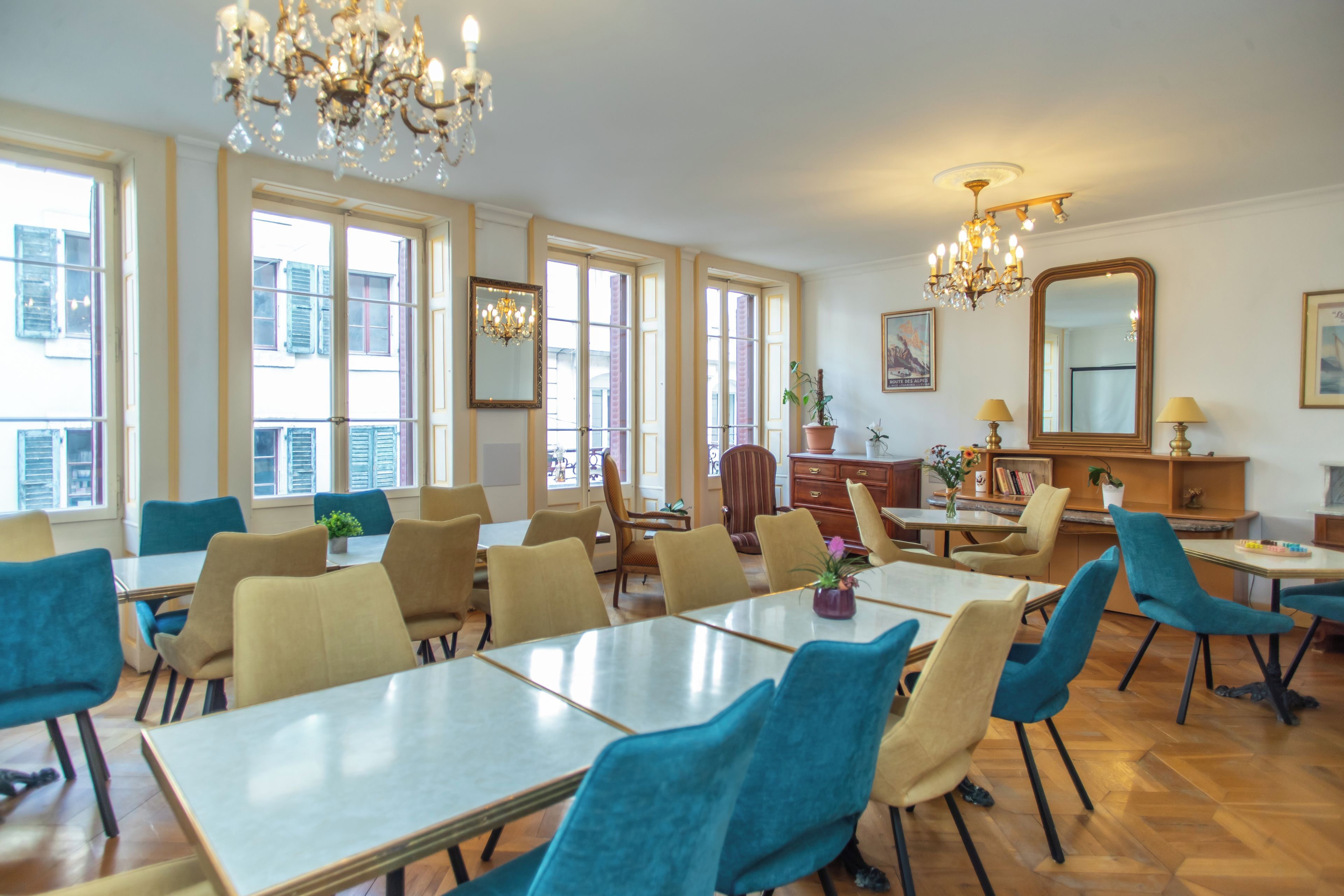 A hotel dining area with multiple tables and chairs, large windows, and chandeliers.