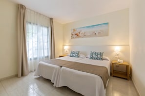 A double bed with a beige bedspread and two pillows, a nightstand with a lamp, and a painting of seashells on the wall. - Hotel THB Royal (Yaiza)