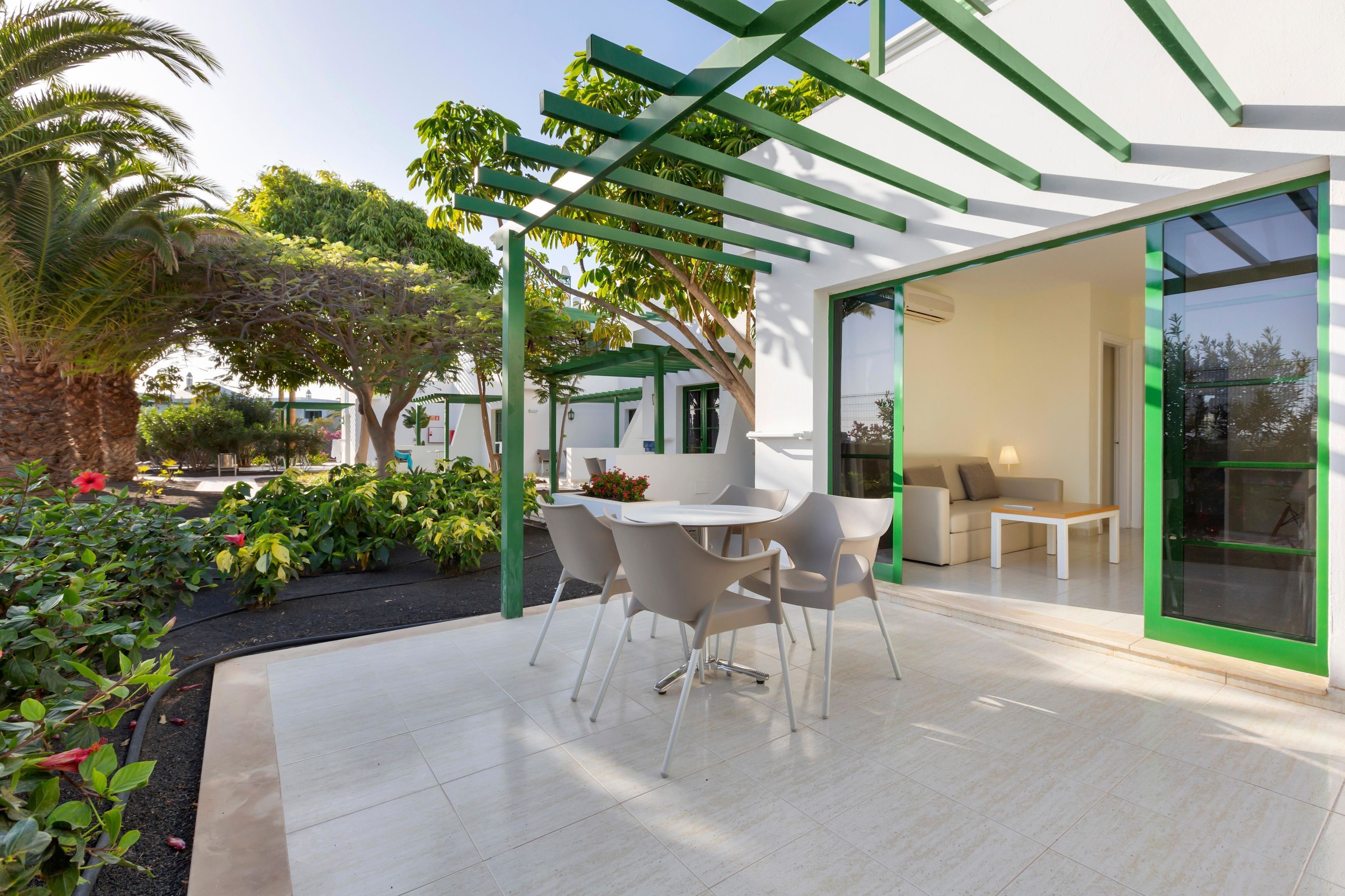 Un patio al aire libre con una mesa y sillas blancas, rodeado de follaje y un diseño arquitectónico moderno.