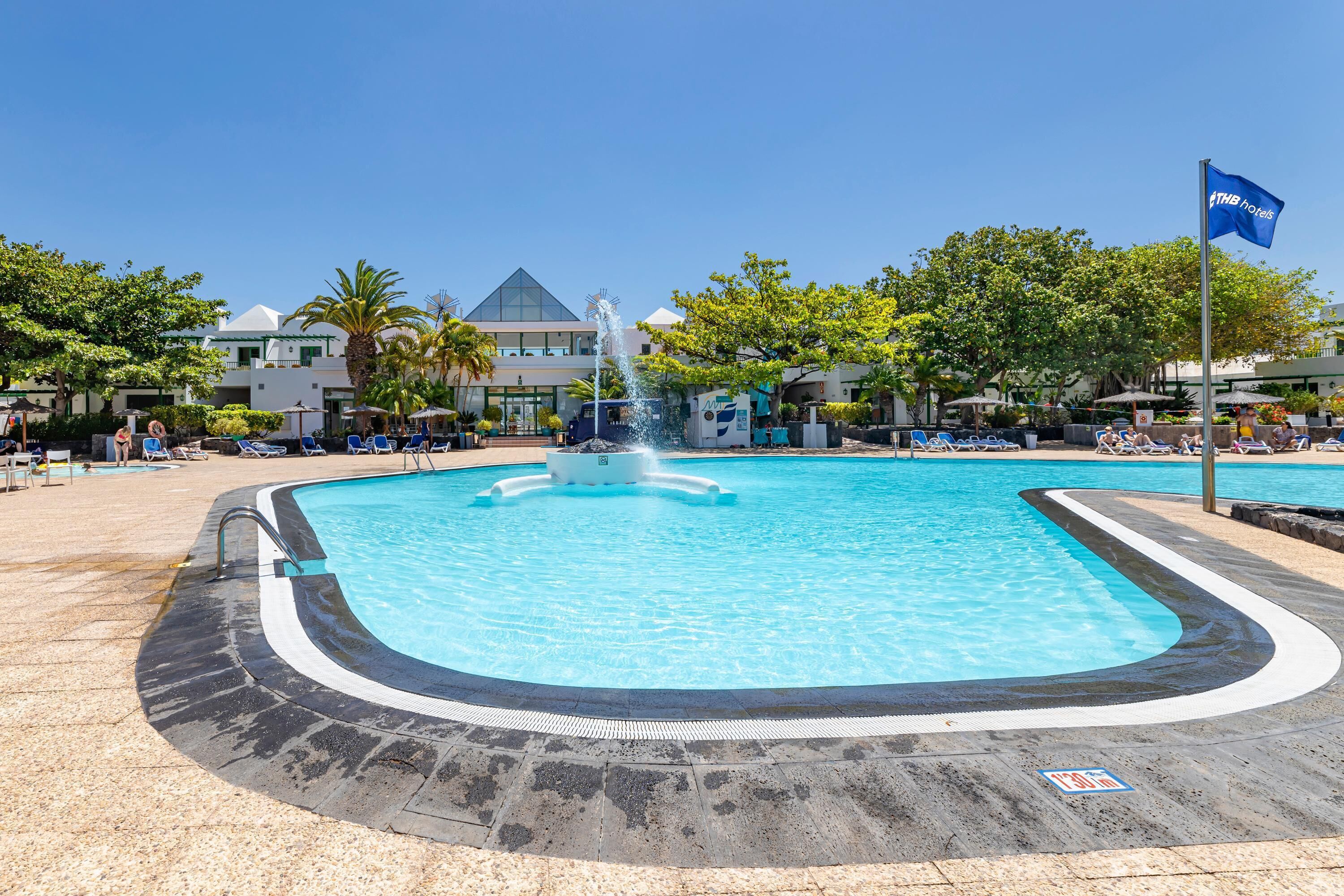 A swimming pool with a fountain, surrounded by lounge chairs and umbrellas, with a hotel building in the background.