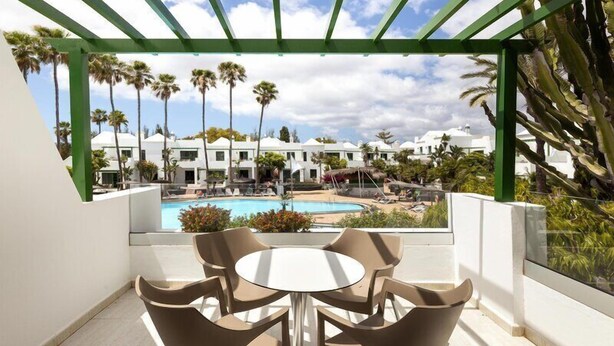 A covered outdoor area with a pool, palm trees, and a view of a resort. - Hotel THB Royal (Yaiza)
