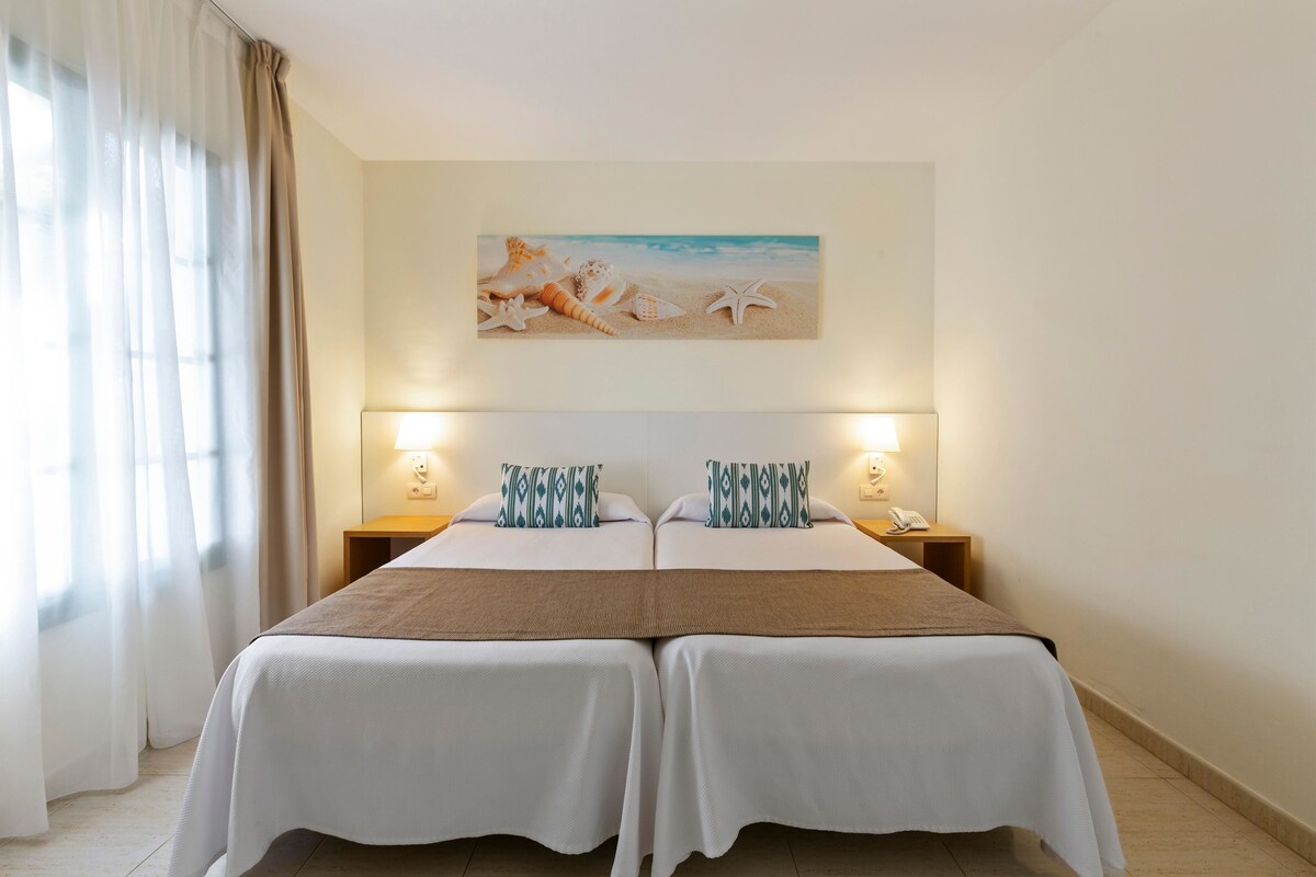A double bed with white and brown bedding, two bedside tables with lamps, and a beach-themed painting on the wall.