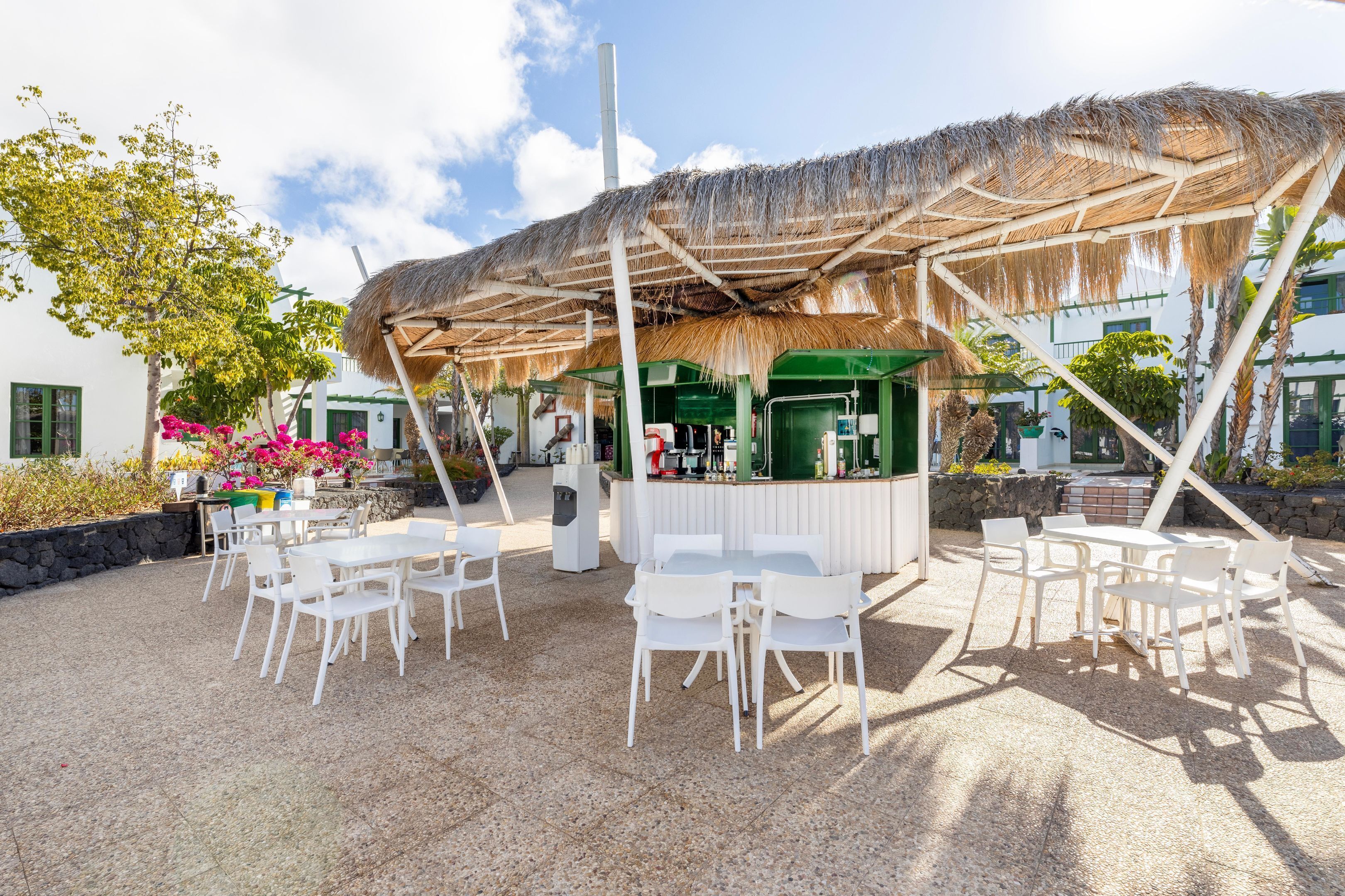 An outdoor seating area with white tables and chairs, a thatched roof structure, and a bar area with a green counter.