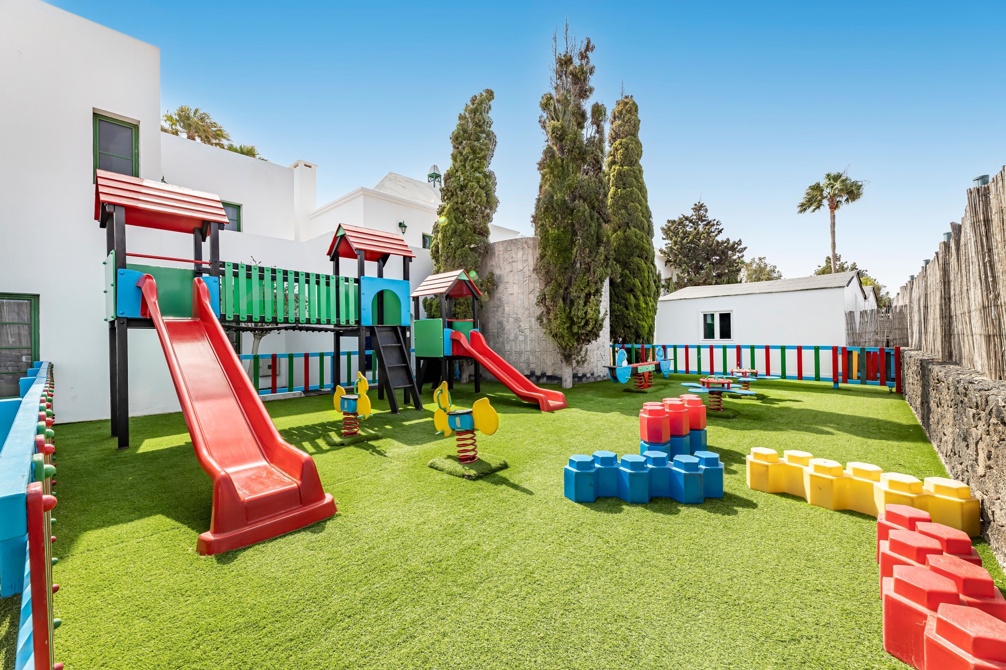 Un parque infantil con toboganes y estructuras para escalar, rodeado por un edificio blanco y árboles altos.