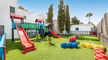 Un parque infantil con toboganes y estructuras para escalar, rodeado por un edificio blanco y árboles altos.