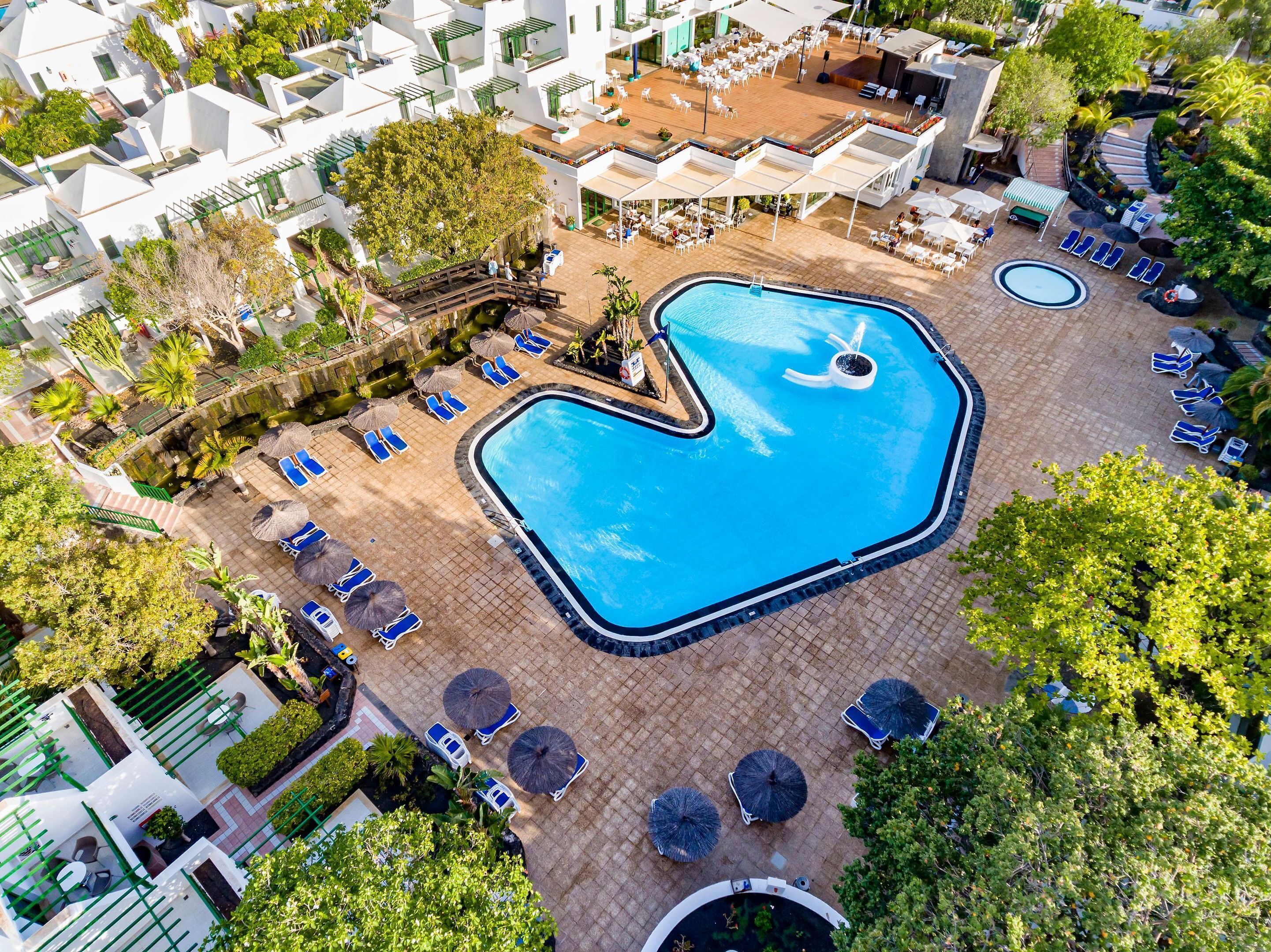 Luchtbeeld van een hotel met een centraal gelegen zwembad, omringd door ligbedden en parasols, plus een overdekt buitengebied met tafels en stoelen.
