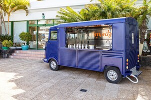 Un camión de comida azul estacionado frente a un edificio con entrada de vidrio y palmeras.