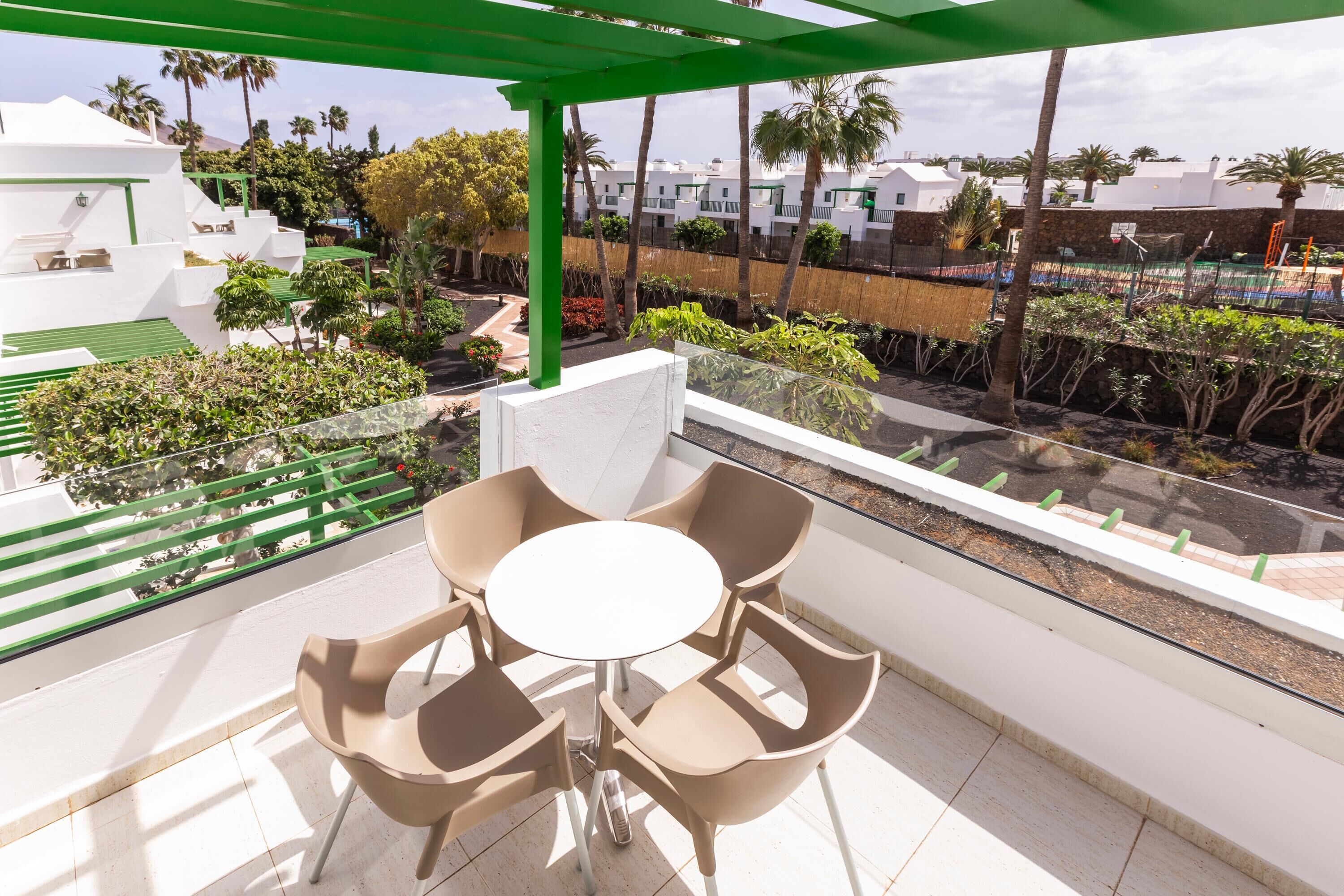 A balcony with a table and chairs, a green pergola, and a view of a residential area with white buildings and palm trees.