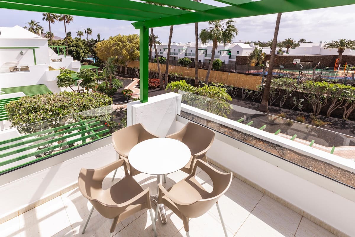 A balcony with a table and chairs, a green pergola, and a view of a residential area with white buildings and palm trees.