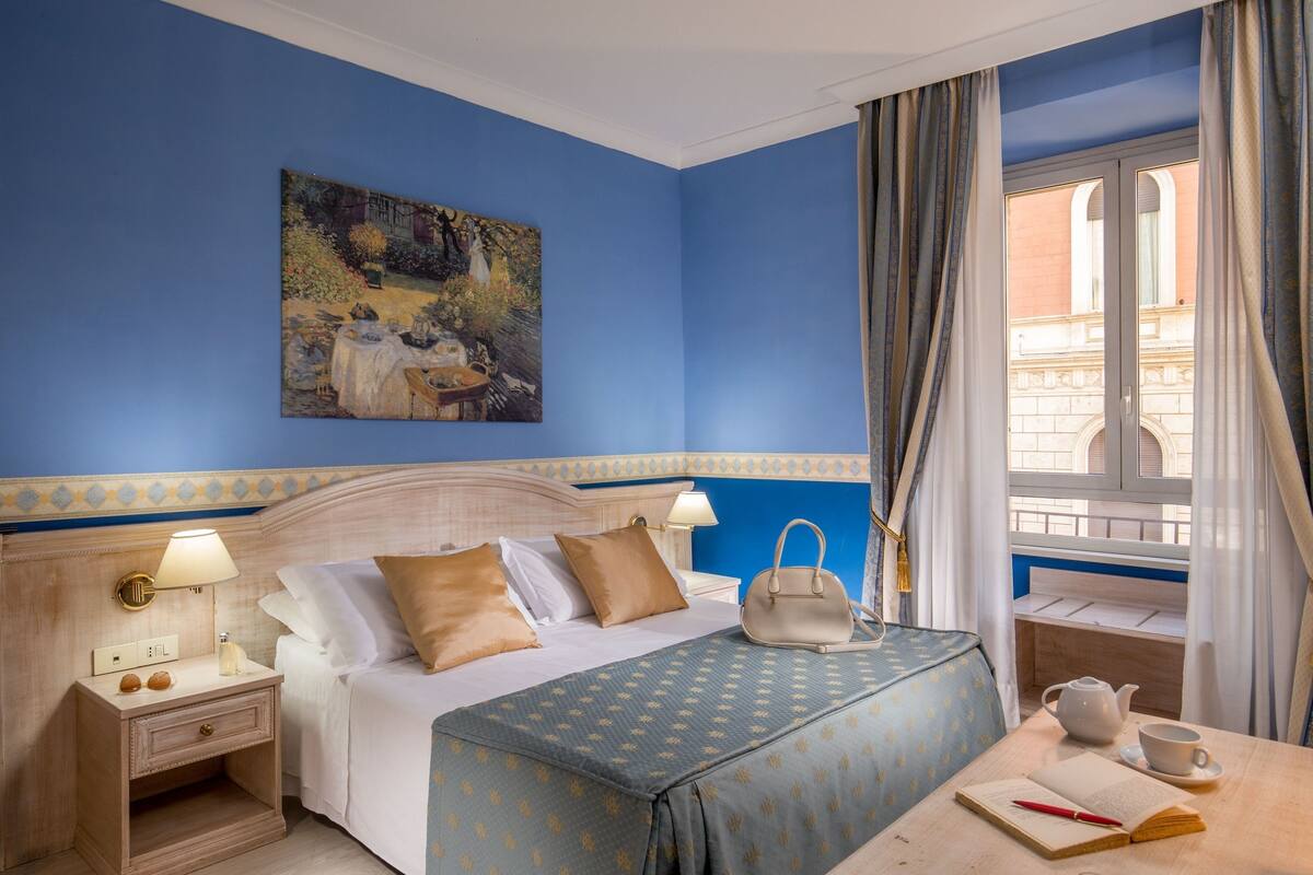 A hotel room with a bed, bedside table, lamp, and a view of the outside.