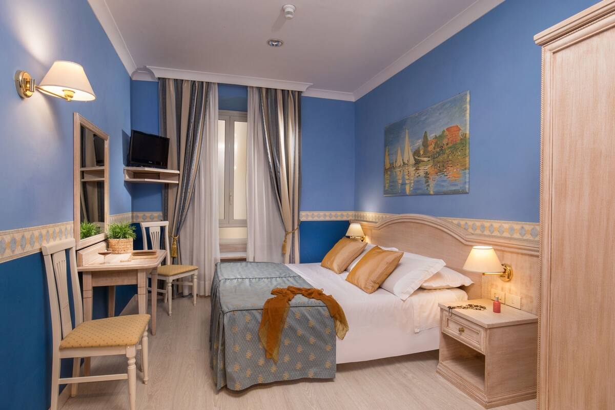 A hotel room with a bed, bedside table, lamp, TV, desk, chair, and curtains.