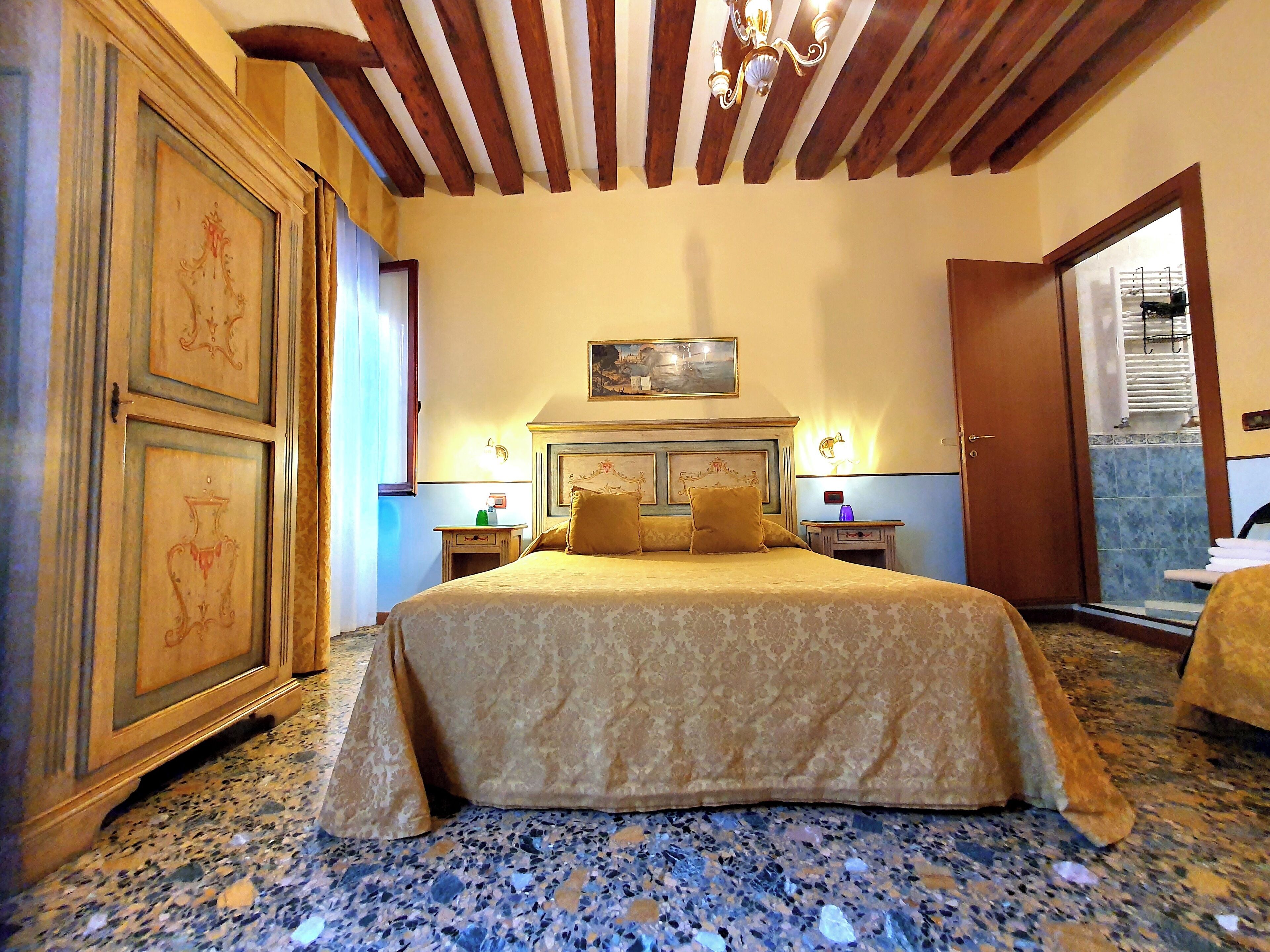 Quarto de hotel com uma cama grande, vigas de madeira no teto e um quadro sobre a cama. Banheiro visível através de uma porta aberta.
