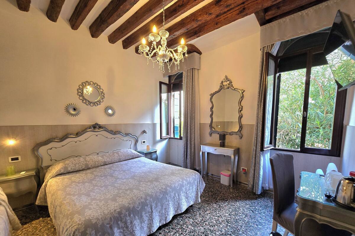 Quarto com cama grande, penteadeira com espelho, lustre e janela com vista para o verde.