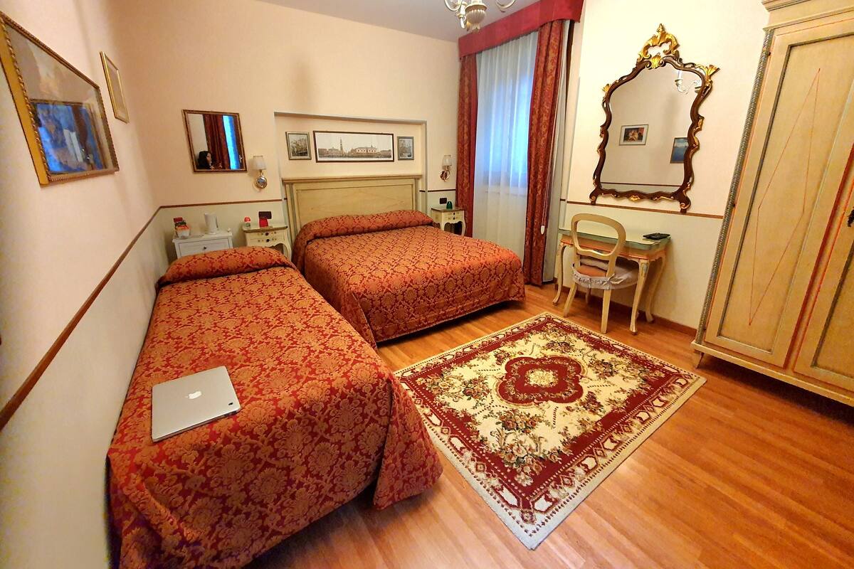 Quarto de hotel com duas camas, uma escrivaninha, uma cadeira, um espelho e um lustre.