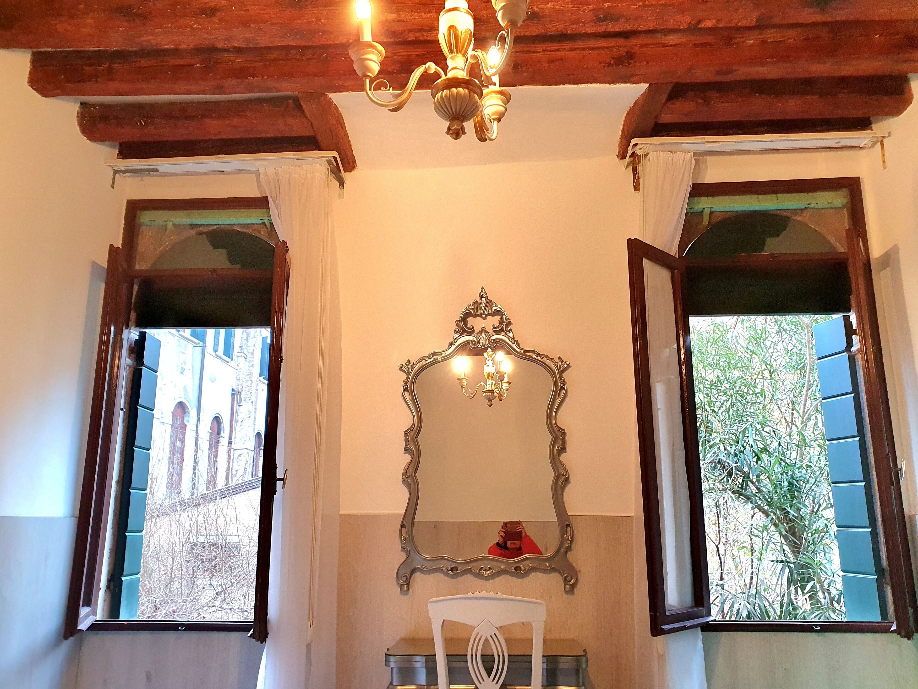 Una stanza con un lampadario, un grande specchio decorato e due finestre con persiane.