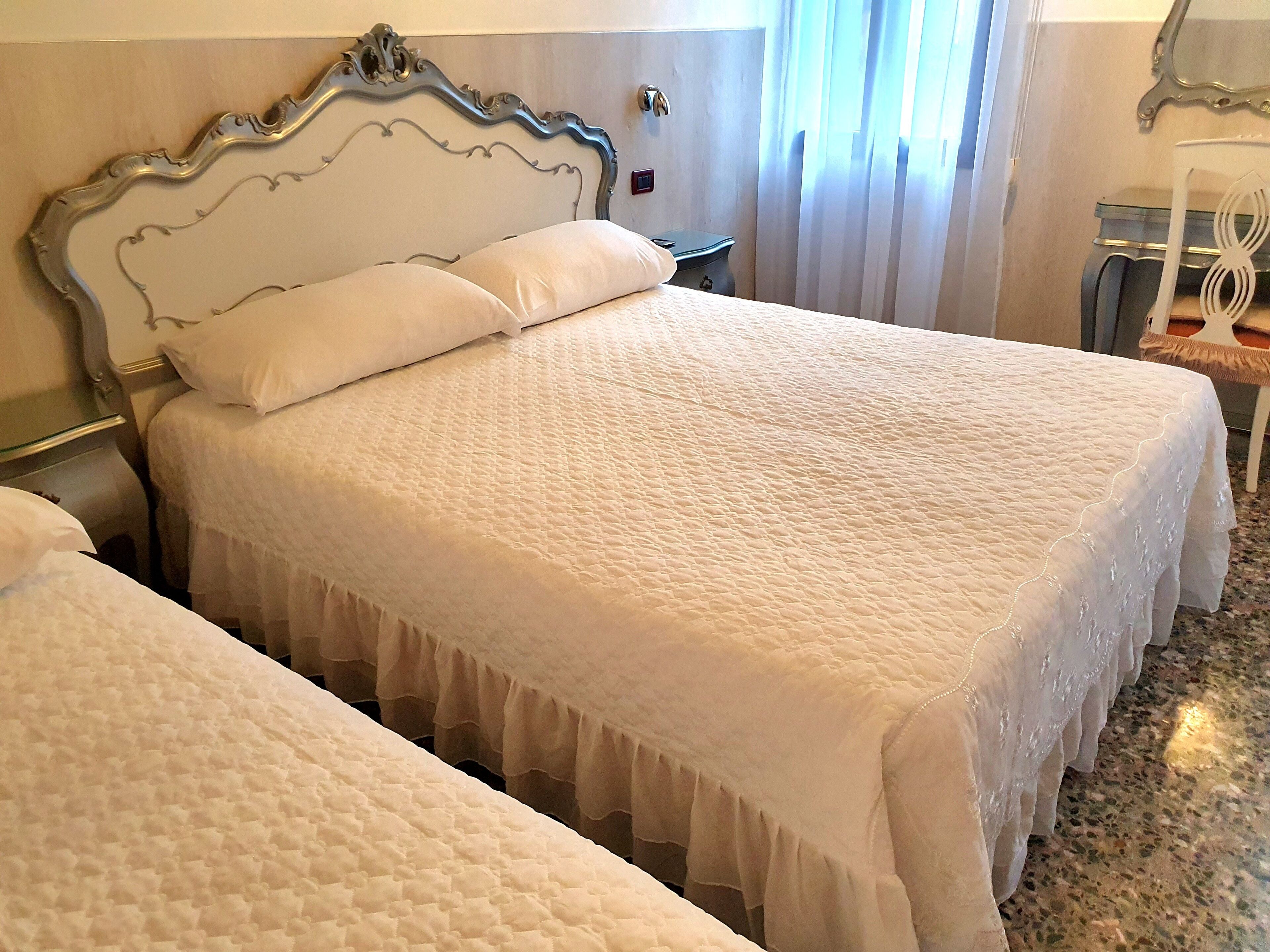 Ein Hotelzimmer mit zwei Betten, jeweils mit weißer, gesteppter Bettdecke und Rüschenvorhang.