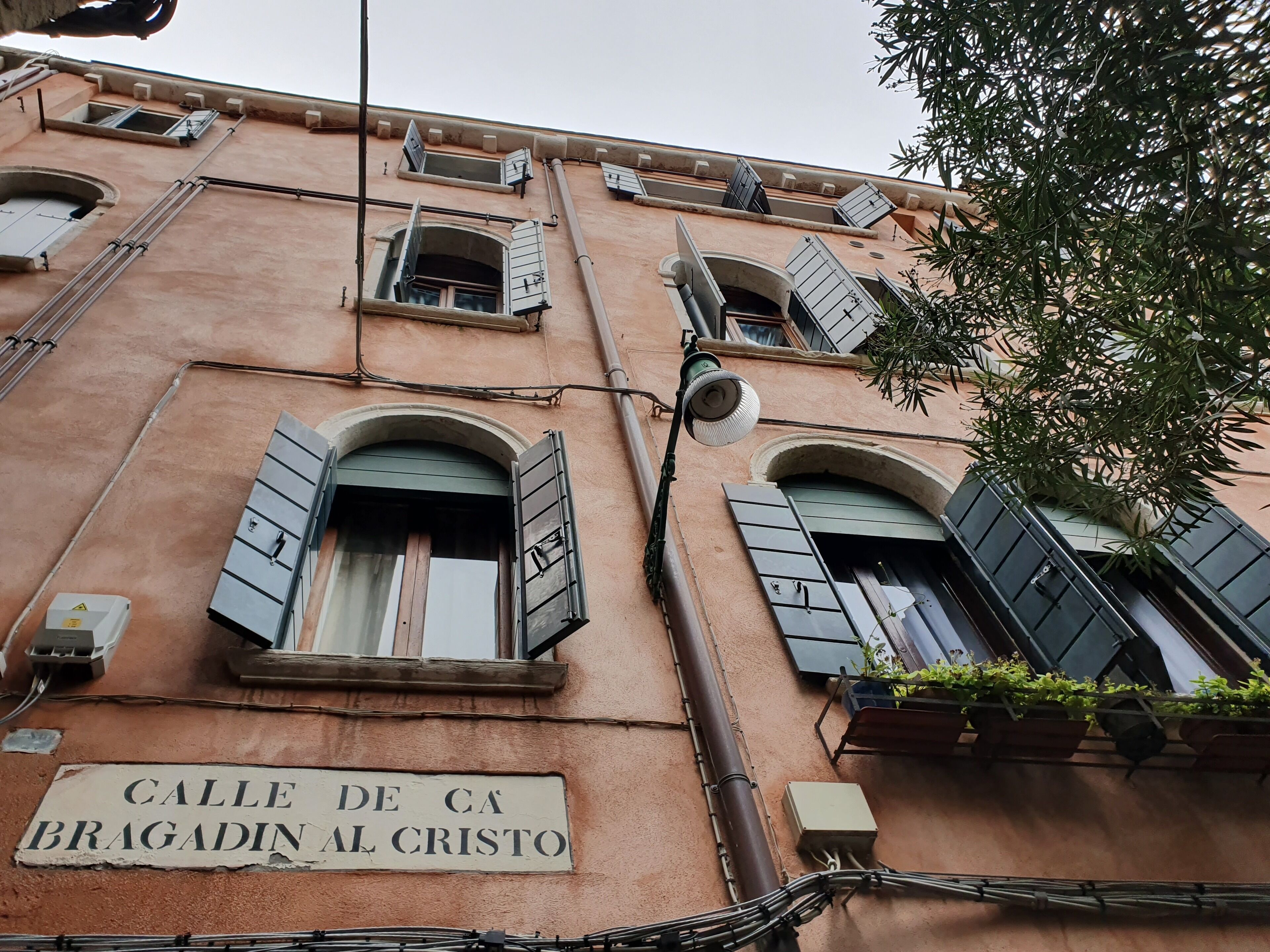 Une façade d’immeuble avec de nombreuses fenêtres, dont certaines sont dotées de volets, et une enseigne indiquant : « Calle de Ca Bragadín al Cristo ».