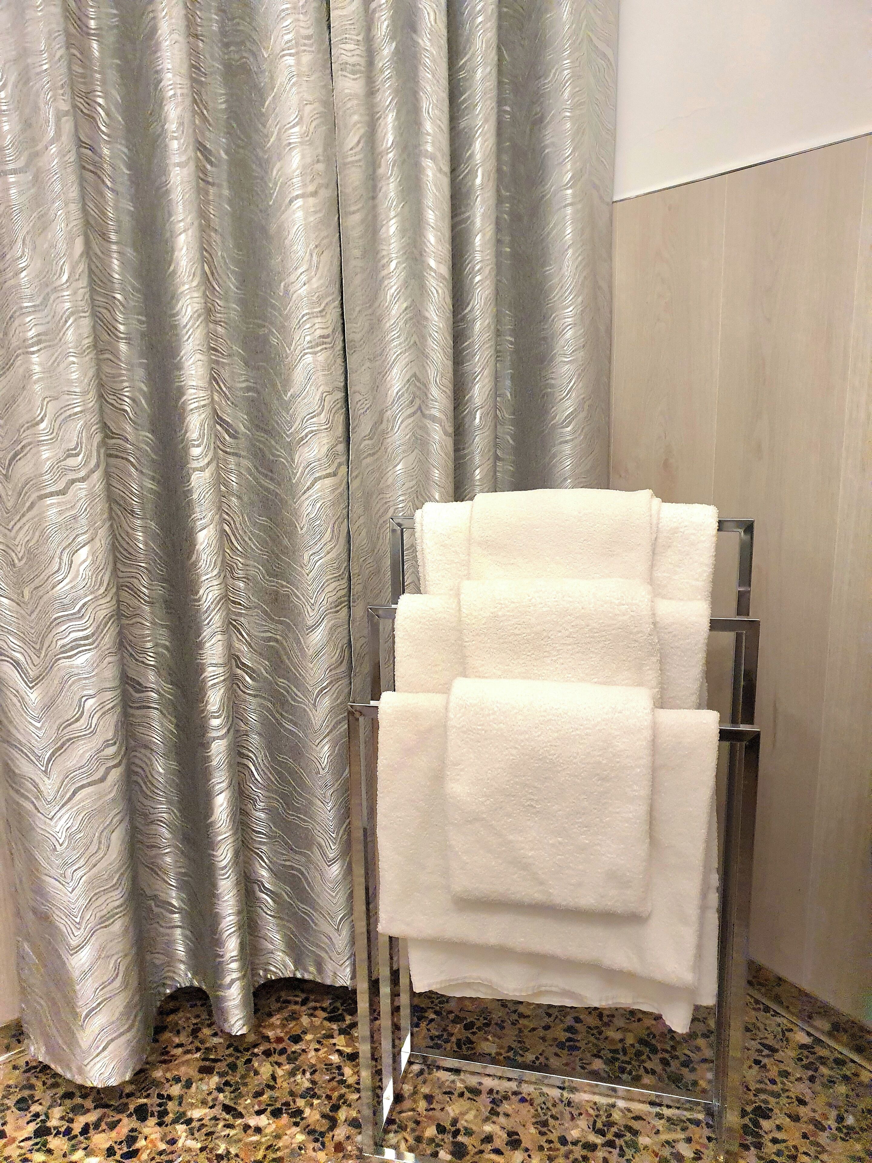 A bathroom with a towel rack holding two white towels, a patterned curtain, and a speckled floor.