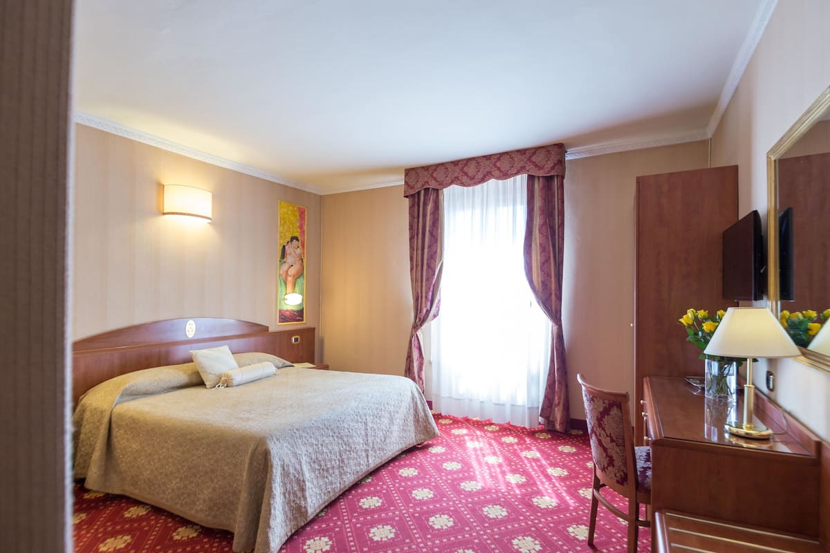A hotel room with a bed, a desk, a chair, a lamp, a painting, and a window with curtains.
