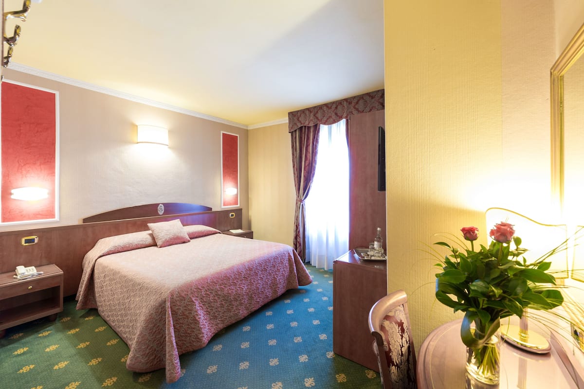 A hotel room with a bed, bedside table, lamp, and a vase with flowers.