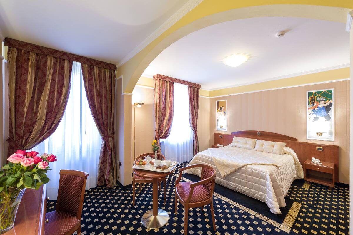 A hotel room with a bed, a dining table with chairs, a vase with flowers, and curtains.
