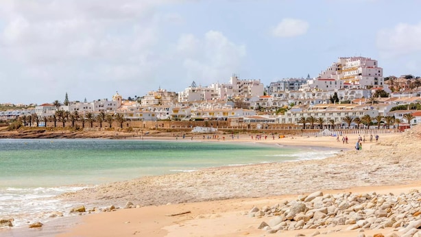 Una playa con un primer plano rocoso, una playa arenosa y un pueblo costero con edificios blancos al fondo.