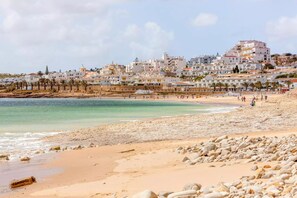 A beach with a rocky foreground, a sandy beach, and a coastal town with white buildings in the background. - Quinta dos Caracóis (Lagos)