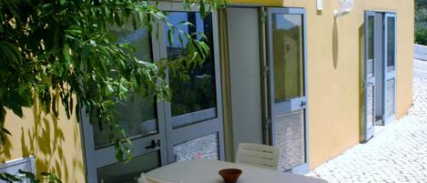 An outdoor patio with a white table and chairs, a bowl on the table, and a yellow building with glass doors and windows.