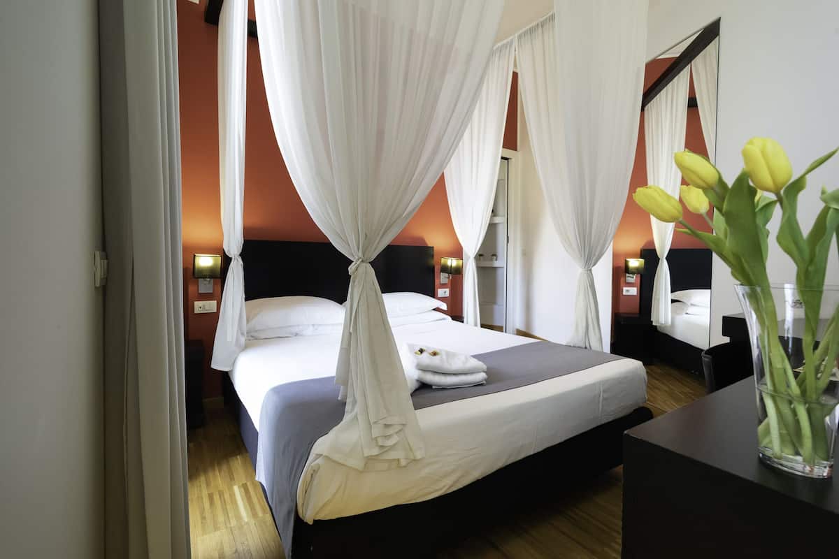 A hotel room with a bed, white curtains, a nightstand, a lamp, and a vase with flowers.