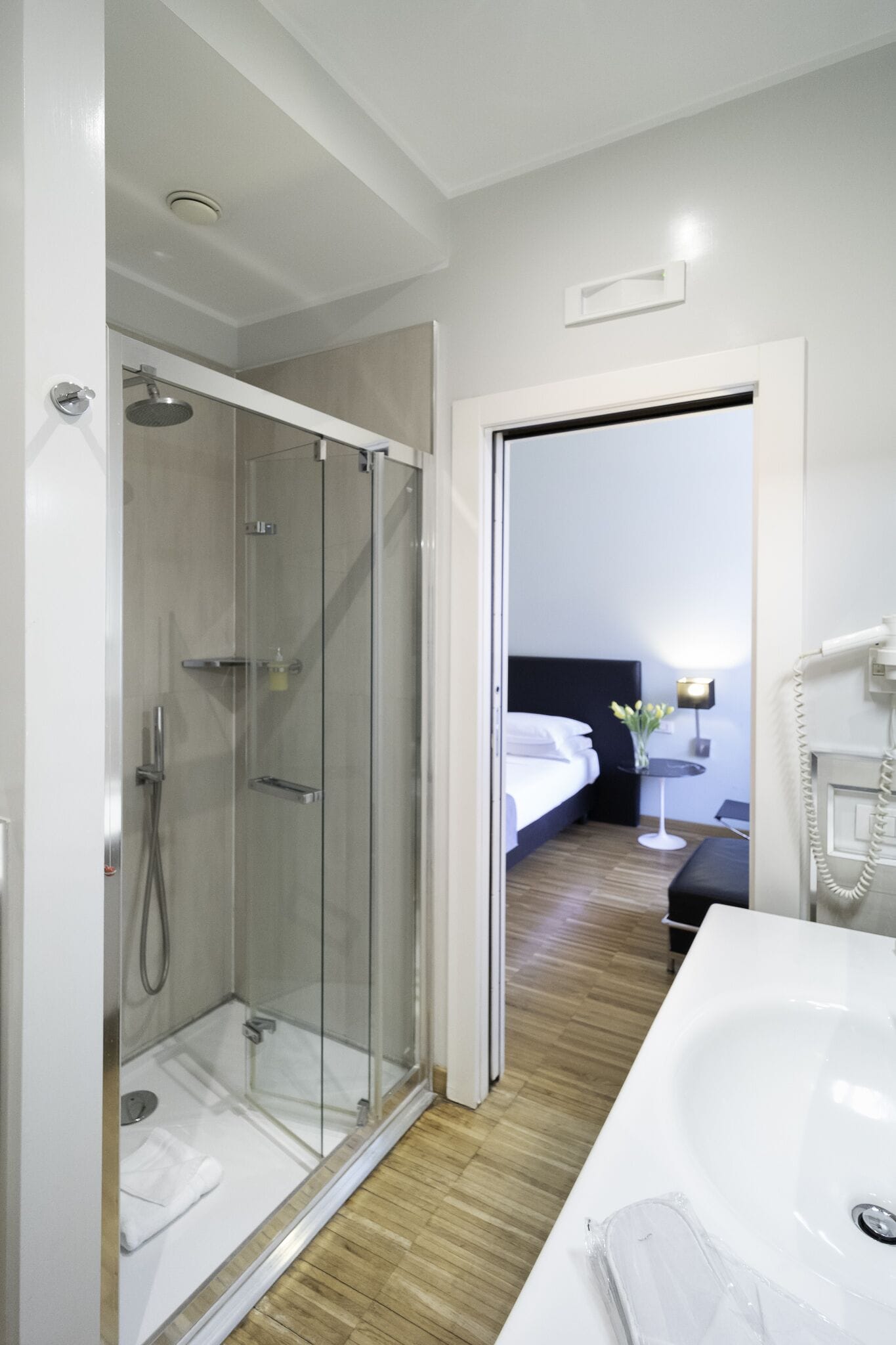 Une chambre d’hôtel moderne dotée d’une douche en verre, d’une grande baignoire blanche, d’un lit parfaitement fait et d’une petite table de chevet avec une lampe.