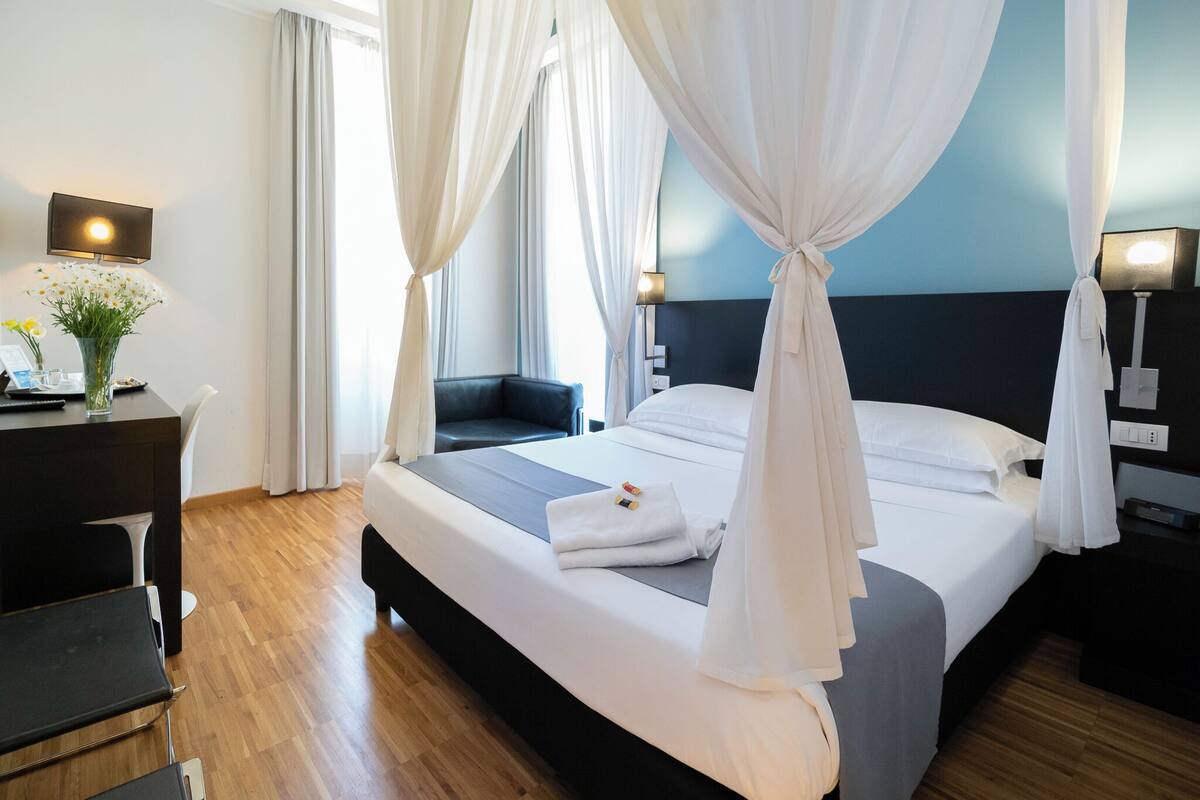 A hotel room with a bed, bedside tables, a desk, a chair, and a window with curtains.