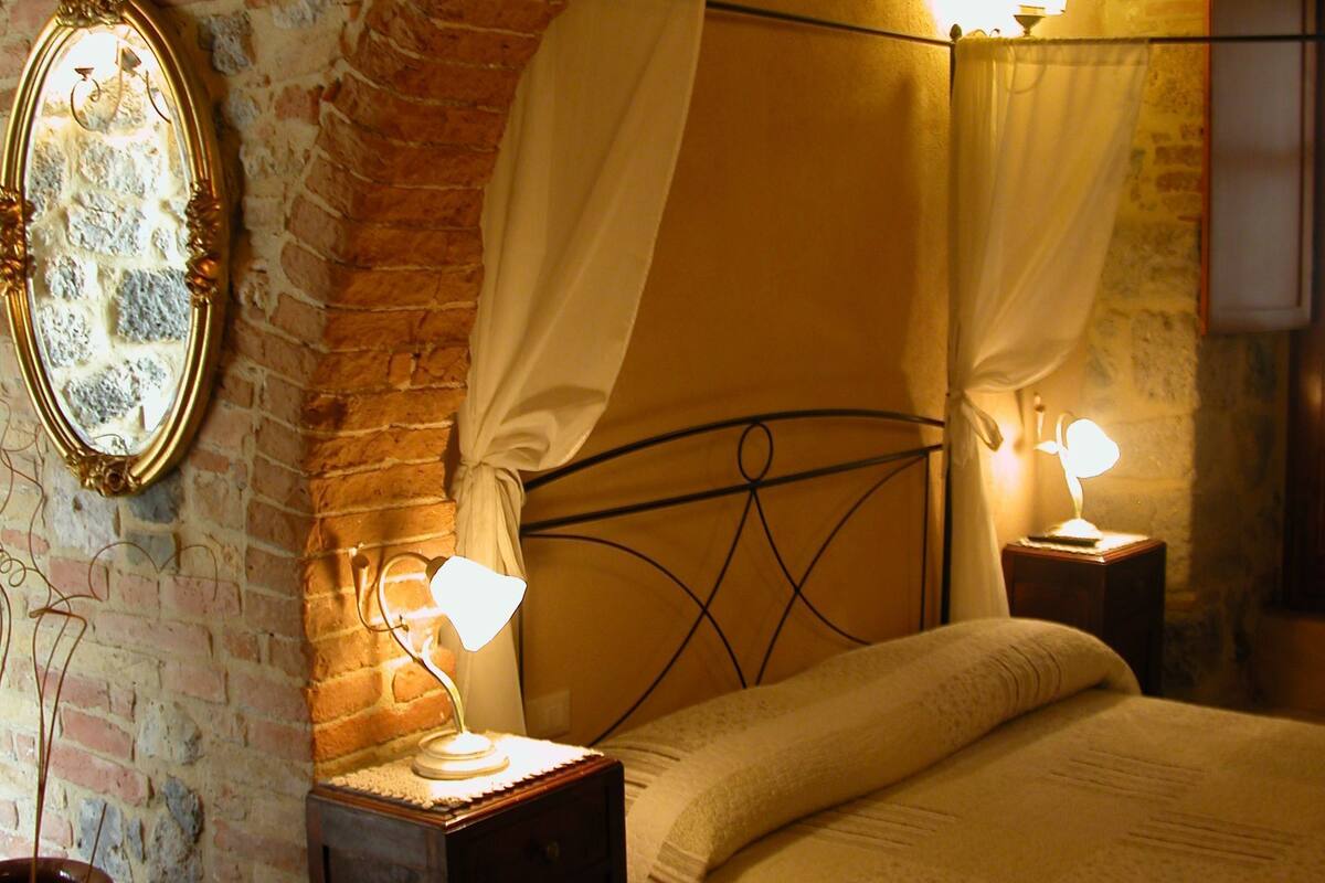 A bedroom with a brick wall, a bed with a canopy, two bedside tables with lamps, and a mirror on the wall.