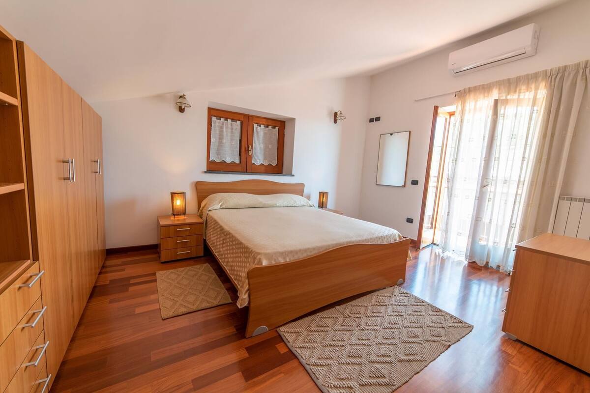 Una camera da letto con un letto di legno, un comodino, un armadio, una finestra con tende e un condizionatore d'aria.