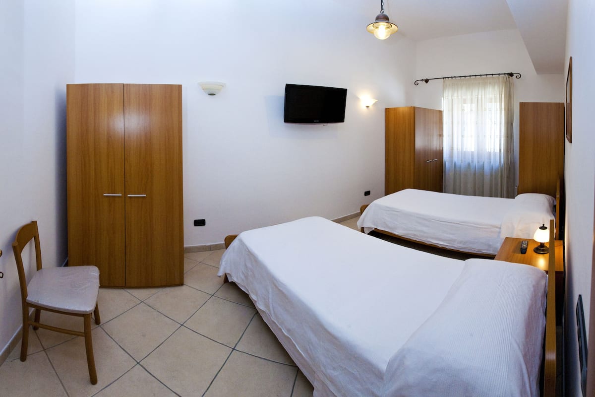 Camera d'albergo con due letti singoli, un armadio in legno, una sedia, una televisione e una finestra con tende.