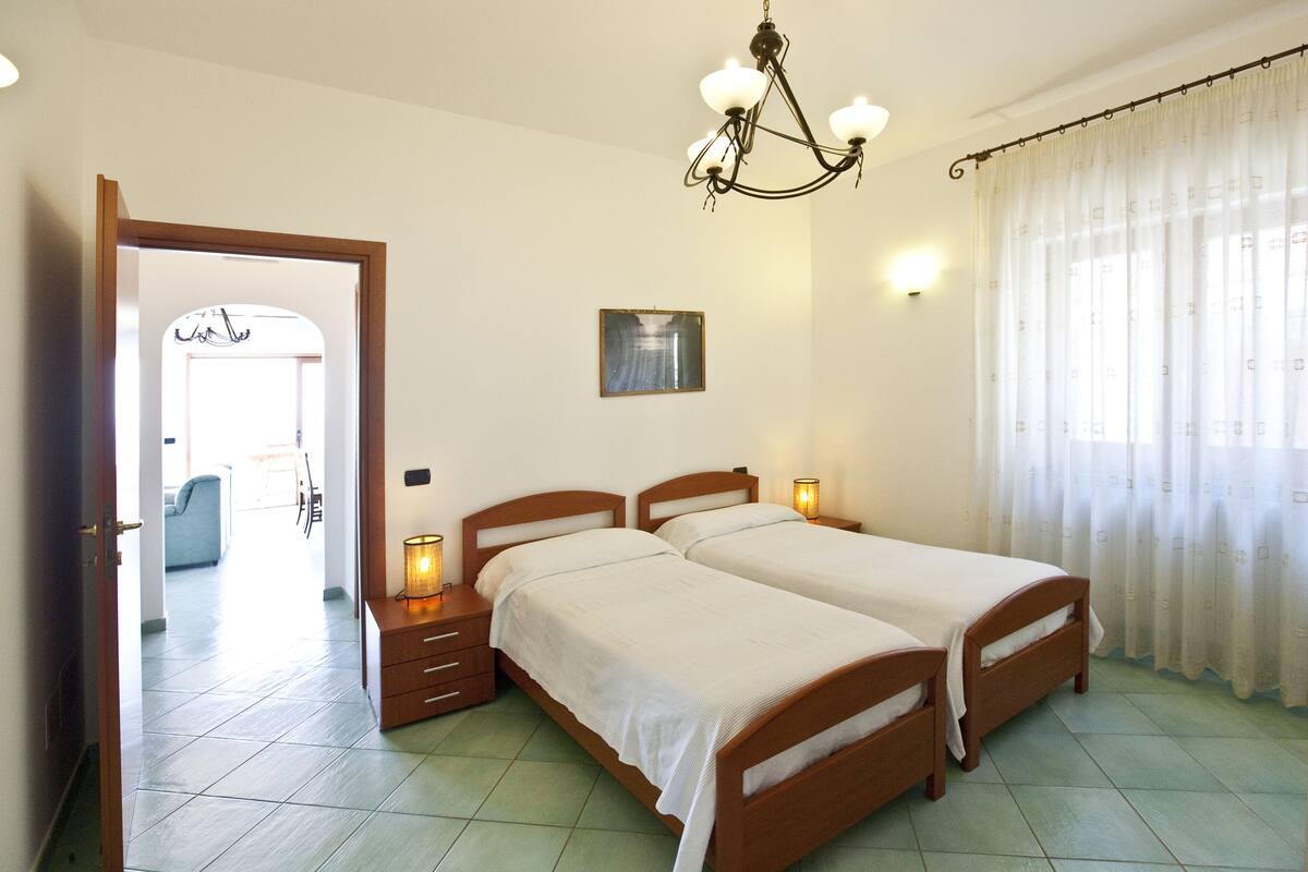 Una camera da letto con due letti, un lampadario, un quadro e vista sull'area soggiorno attraverso una porta aperta.
