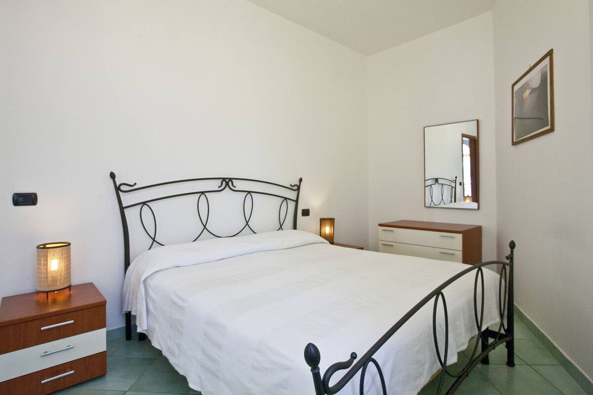 Una camera da letto con struttura letto in metallo, biancheria da letto bianca, comodino con lampada, cassettiera e uno specchio a parete.