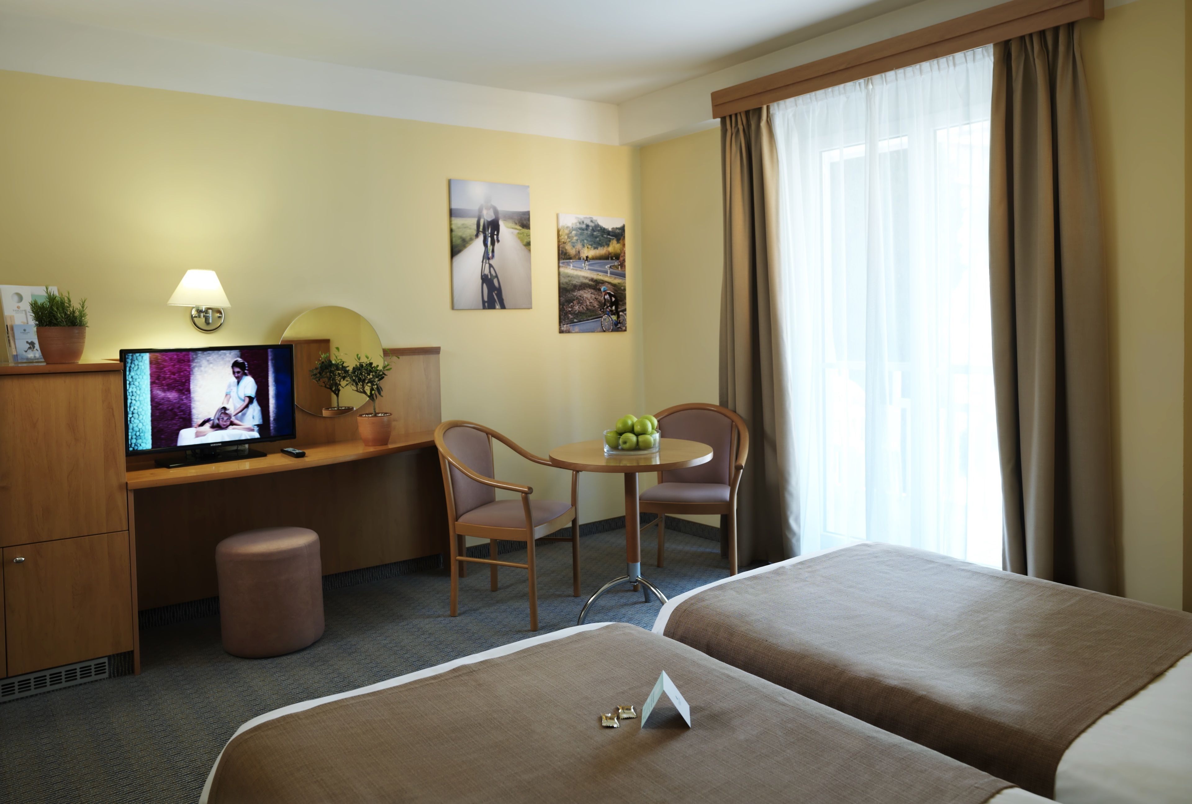 A hotel room with two beds, a desk with a TV, a chair, a small table with apples, and a window with curtains.