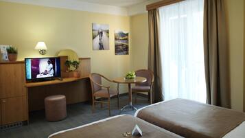 A hotel room with two beds, a desk with a TV, a chair, a small table with apples, and a window with curtains.