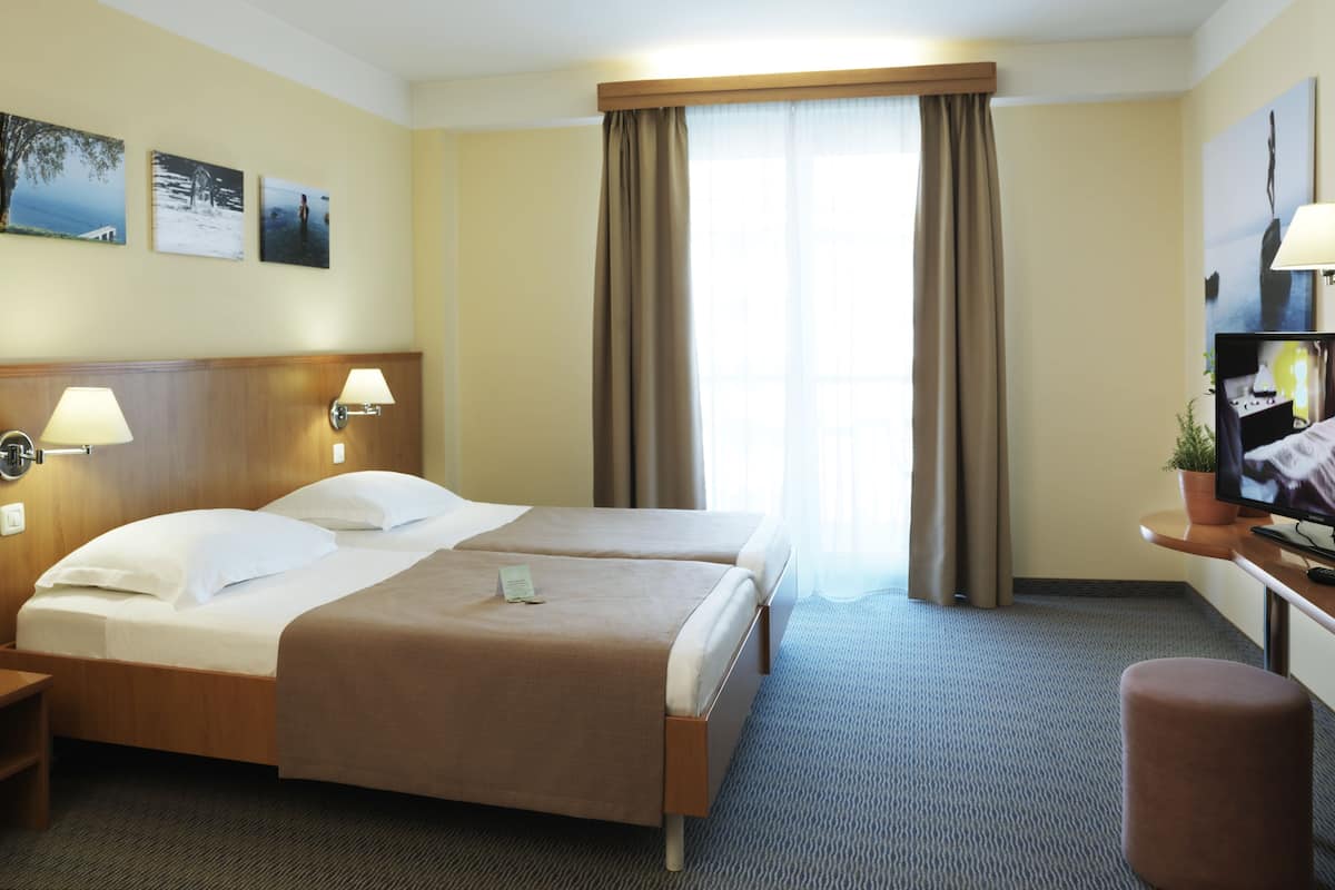A hotel room with two beds, a desk, a TV, and a window with curtains.