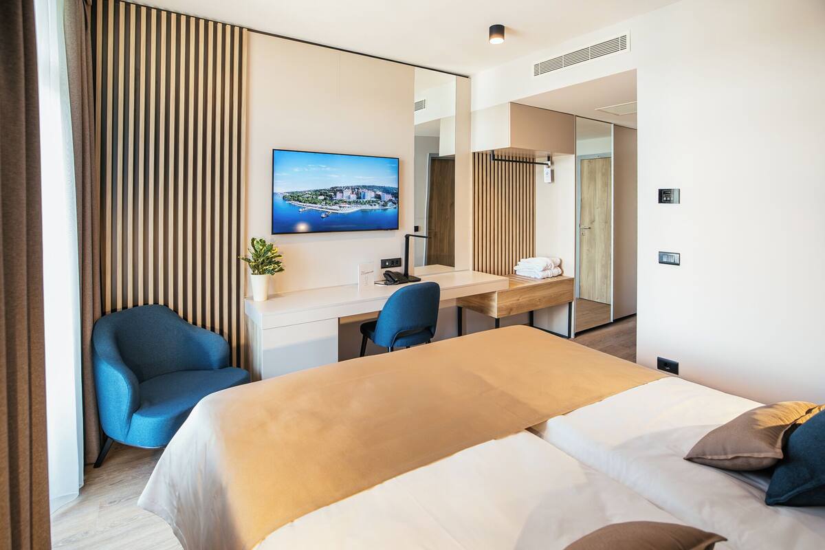 A modern hotel room with a bed, a desk, a chair, and a large window with a cityscape view.