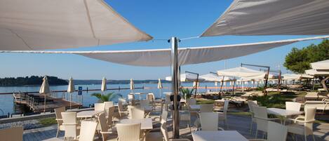 An outdoor seating area with white wicker furniture and large white umbrellas, overlooking a body of water.