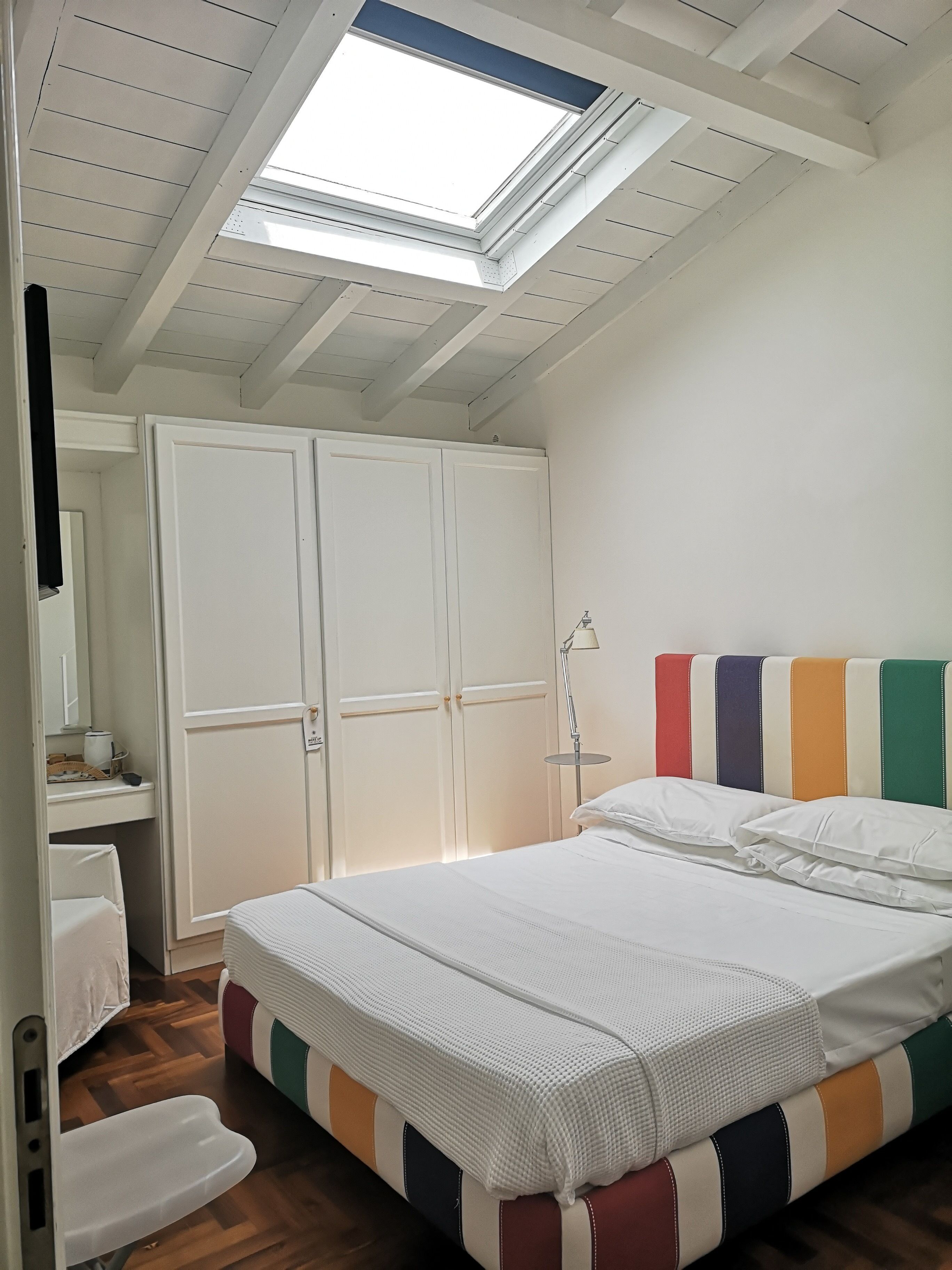 Ein Schlafzimmer mit Dachfenster, einem Bett mit farbigem Kopfteil, einem Nachttisch und einem Kleiderschrank.