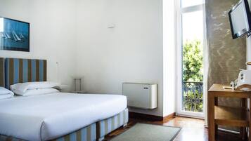 Habitación de hotel con cama, una mesita, un radiador y ventana con vista a follaje.
