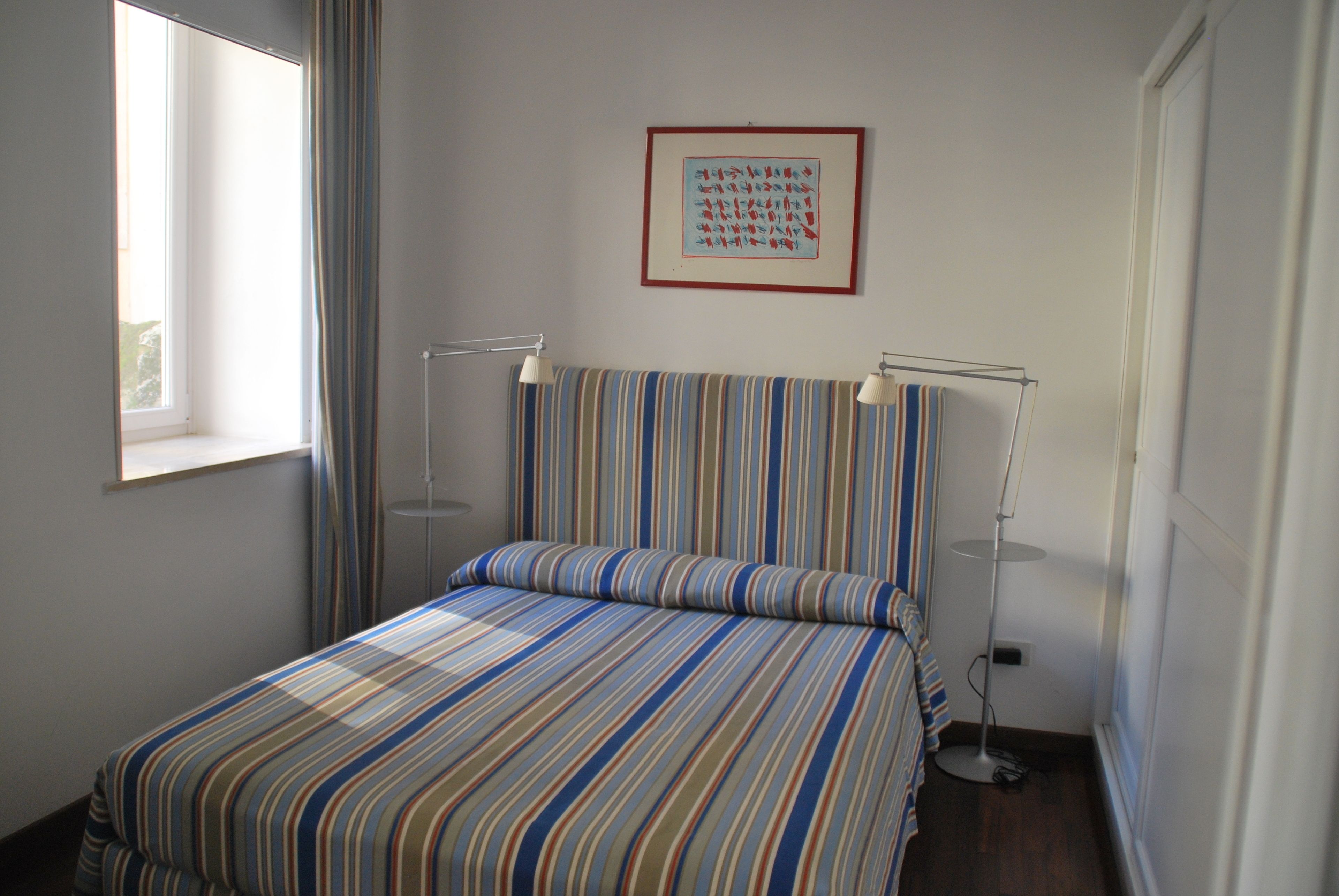 A room with a bed, a window, a framed picture on the wall, and two floor lamps.