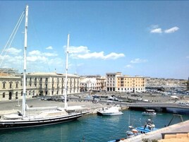 A harbor with a large sailing ship docked, buildings with classical architecture, and a clear blue sky.