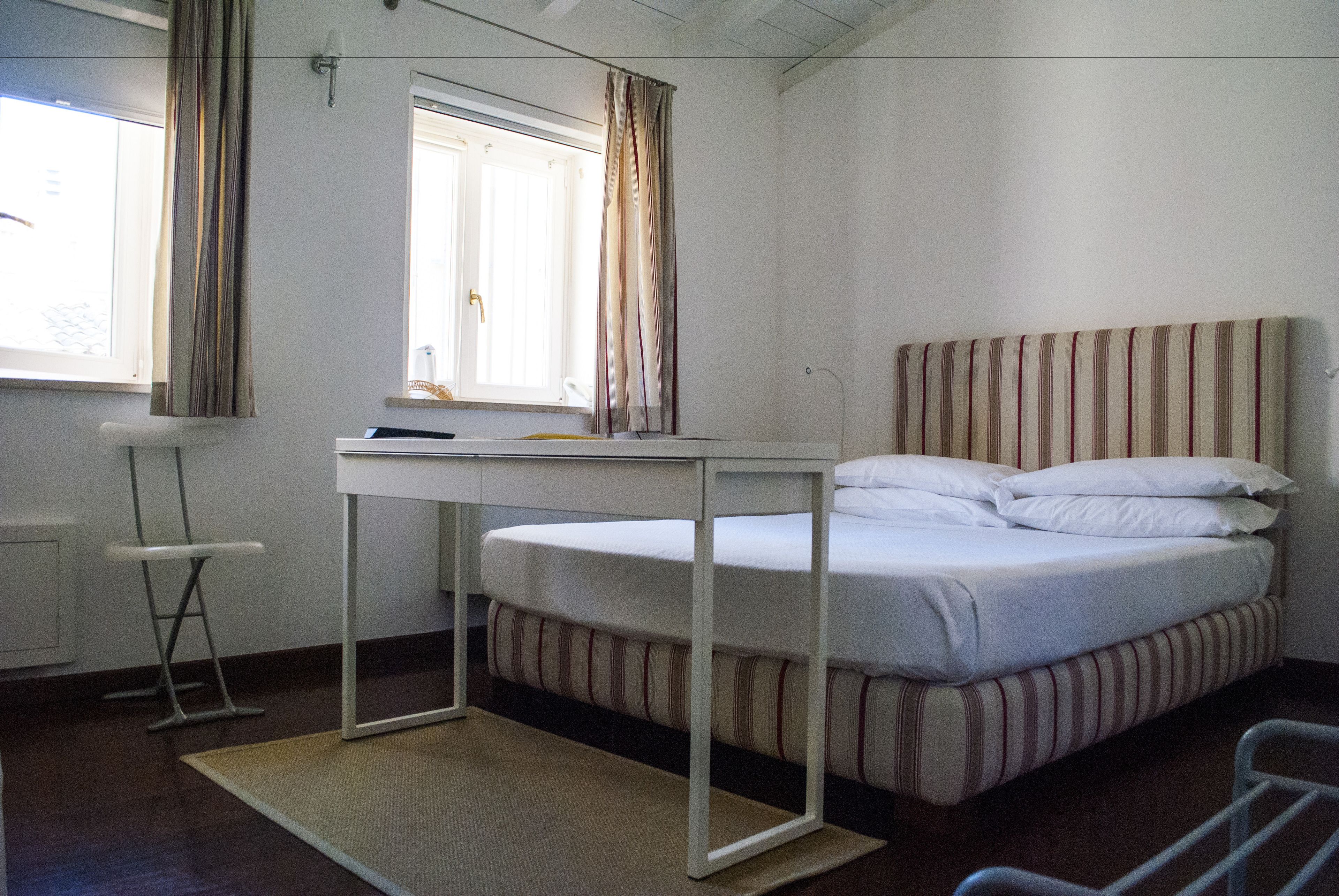 Une chambre individuelle avec un lit simple, un bureau, une lampe sur pied et une fenêtre avec des rideaux.