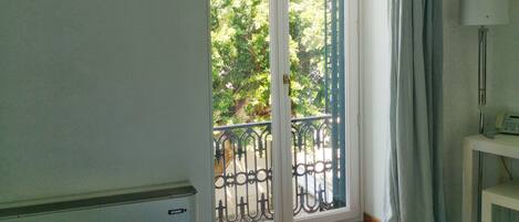 A room with a bed, a window with a view of trees, a balcony with a railing, and a lamp on a side table.