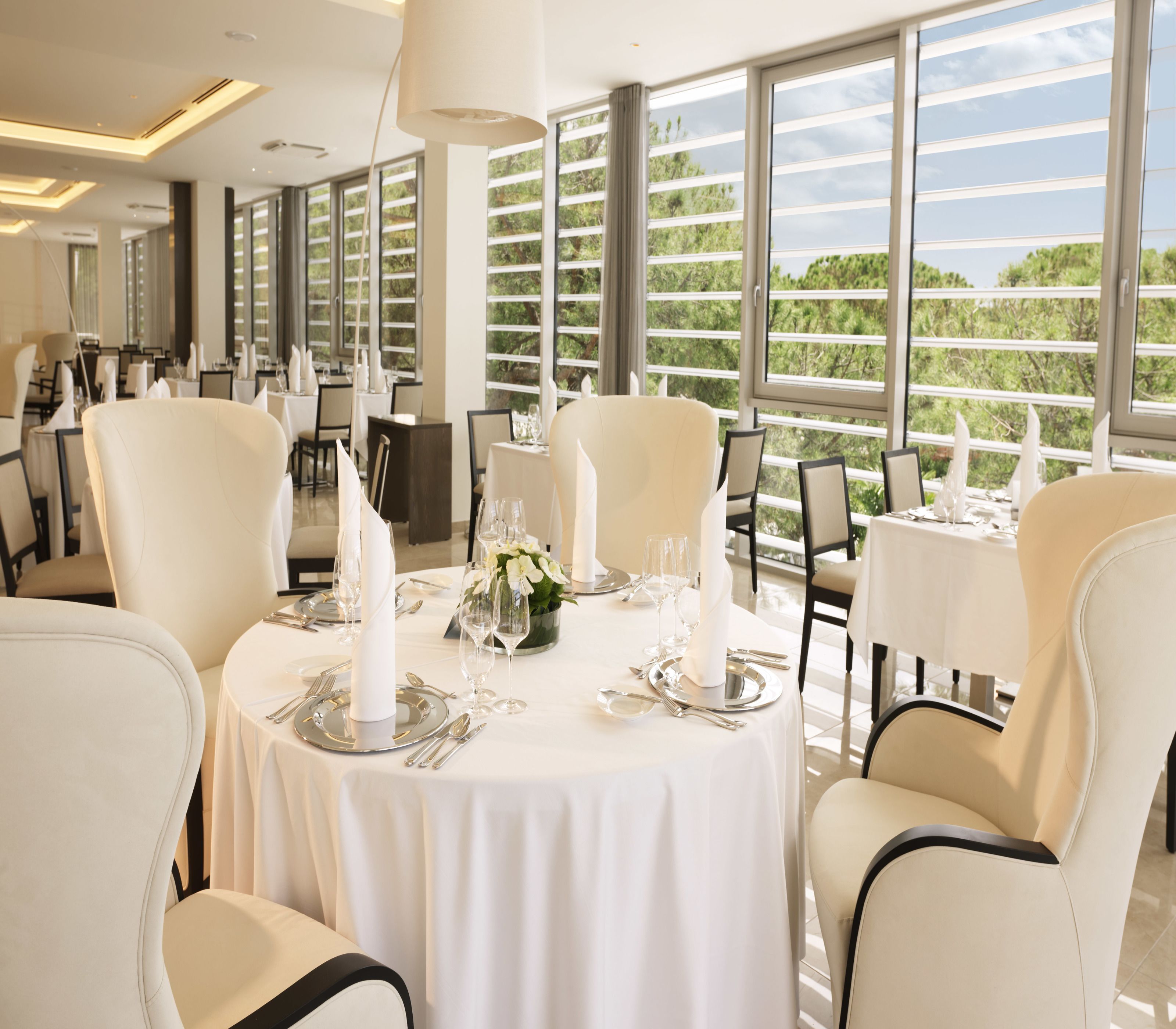 A dining area with round tables set for a meal, featuring white tablecloths, silverware, and candles. Large windows provide a view of greenery outside.